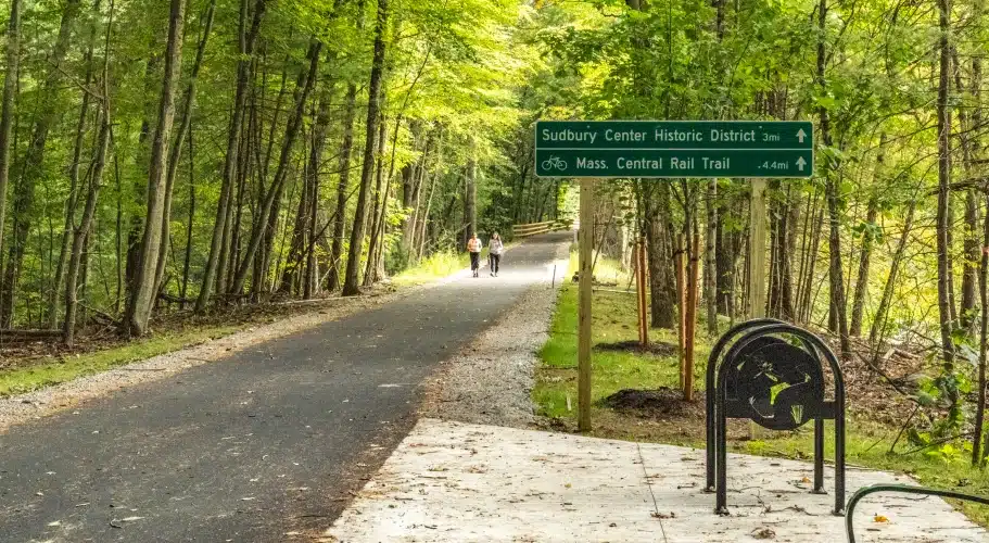 Tree-lined paved trail entrance with directional signs for Sudbury Center and rail trail