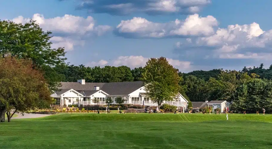 Large white golf clubhouse set against green fairway, trees, and partly cloudy sky