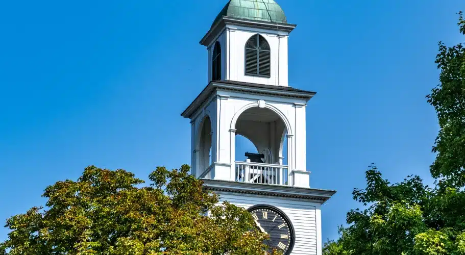 White historic clock tower with green dome against bright blue sky and leafy trees