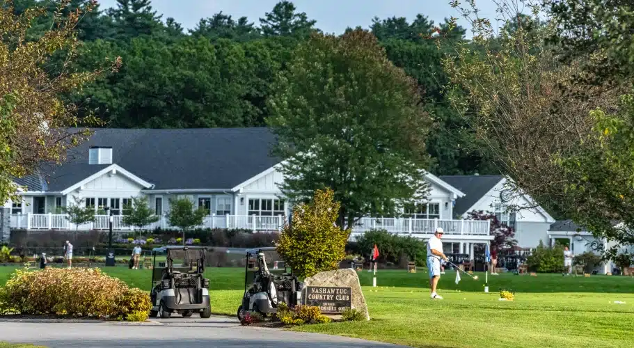 Golfers practice near Nashawtuc Country Club sign, golf carts, and large white clubhouse