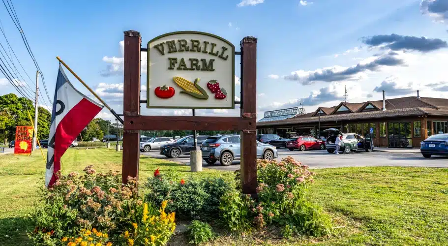Verrill Farm roadside sign surrounded by colorful flowers, parked cars, and blue sky backdrop