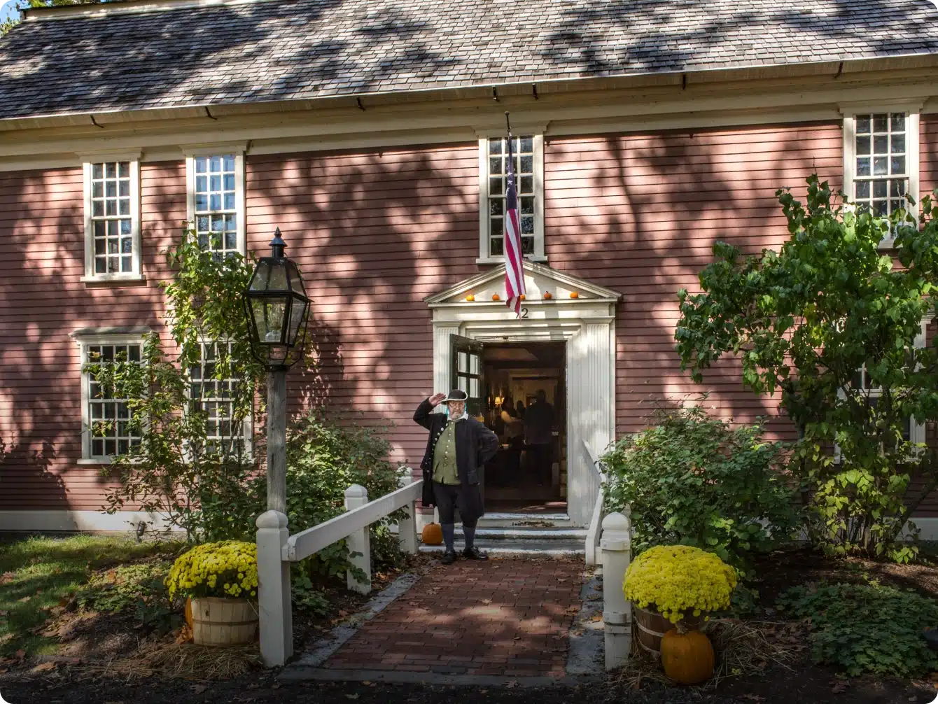 Man in colonial attire greets visitors outside red historic house with fall decor
