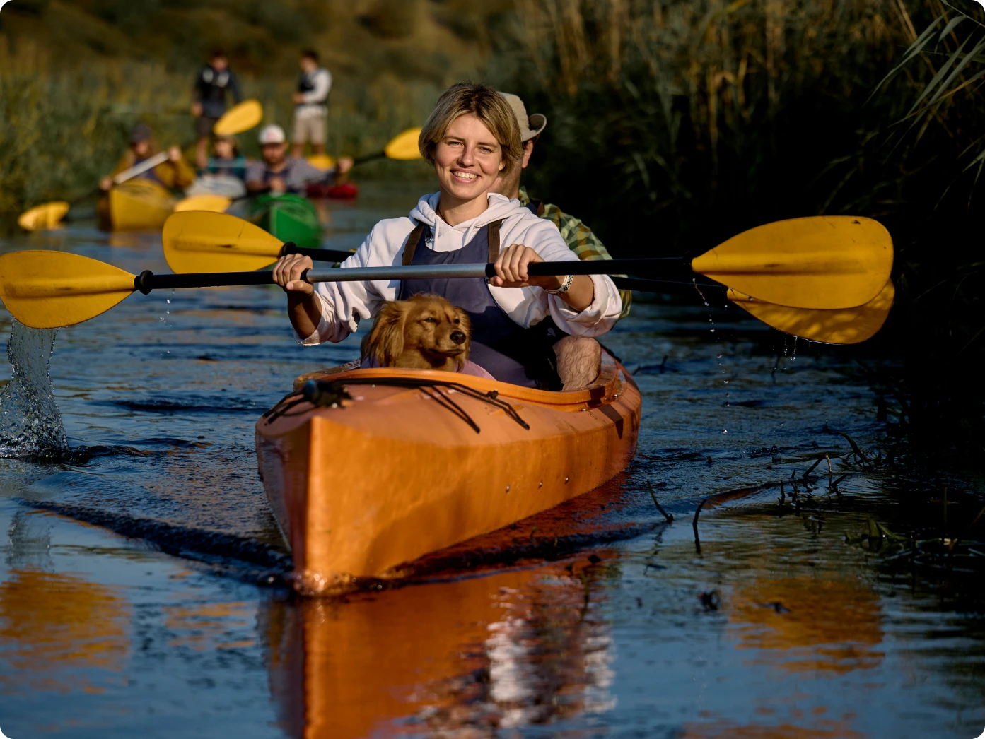 Woman kayaking on calm water with golden retriever dog inside orange kayak at sunset