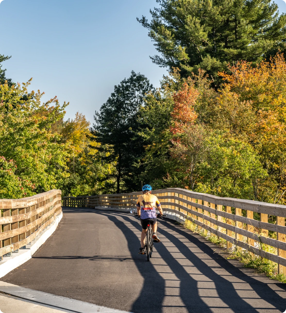Cyclist riding through tree-lined trail with wooden railing and autumn colors