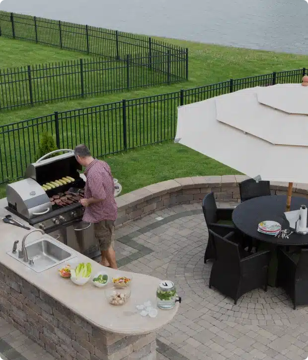 Man grilling vegetables and burgers on built-in patio barbecue beside lake and umbrella seating